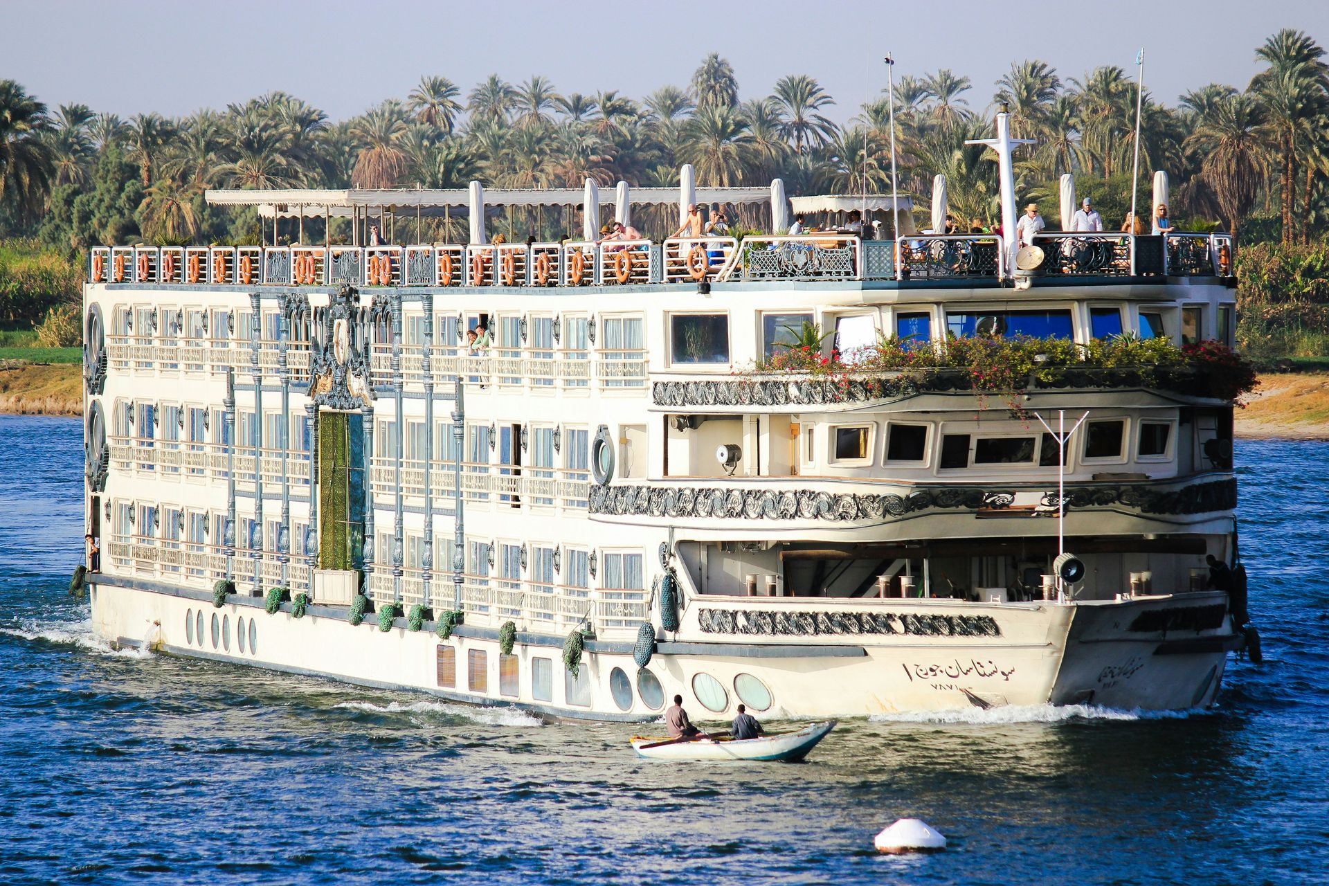 A large white luxury cruise ship, the MS Grand Rose, sailing on the Nile River in Egypt at sunset with desert mountains and palm trees in the background.